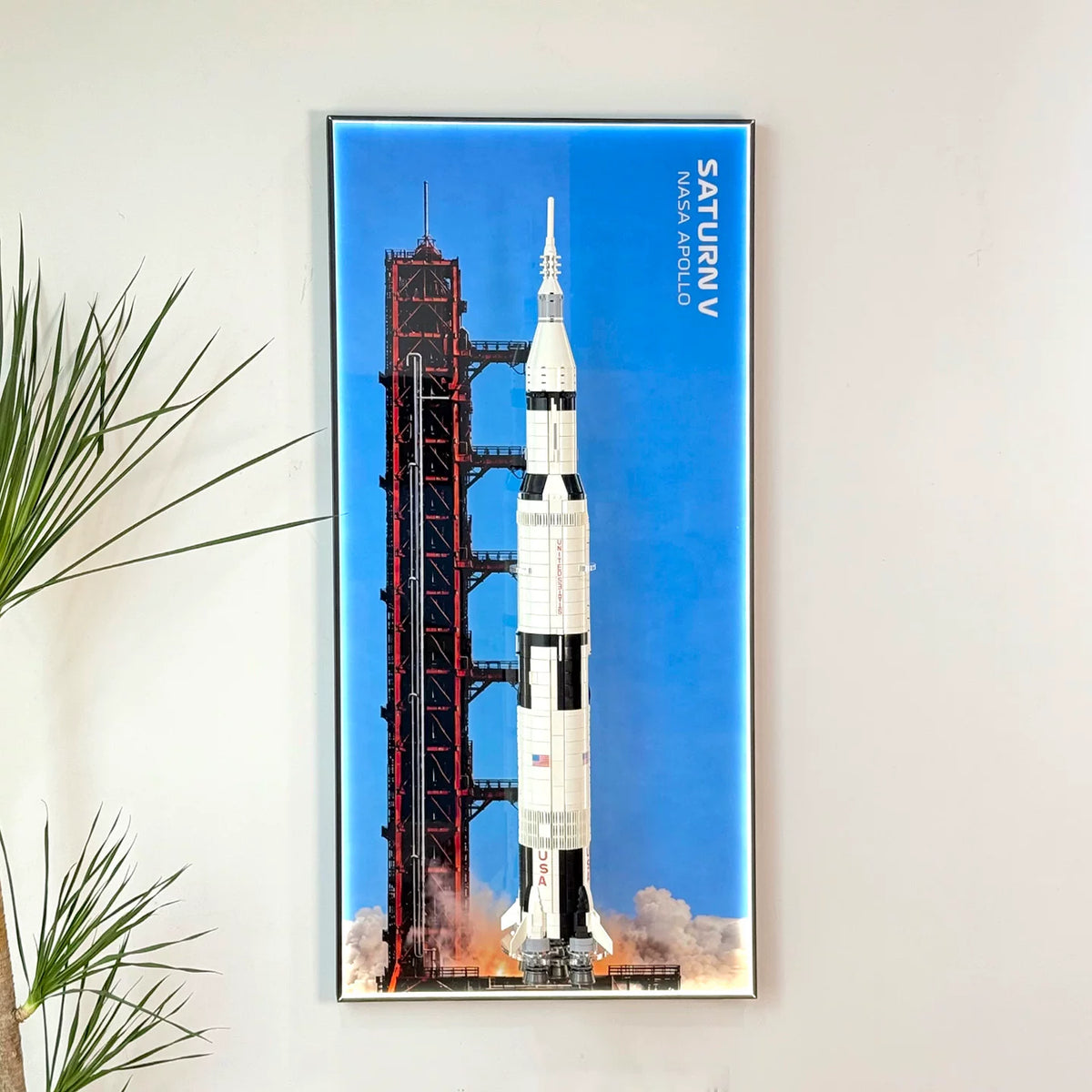 Wall-mounted display frame for LEGO® NASA Apollo Saturn V (21309), shown with rocket model and blue launch backdrop